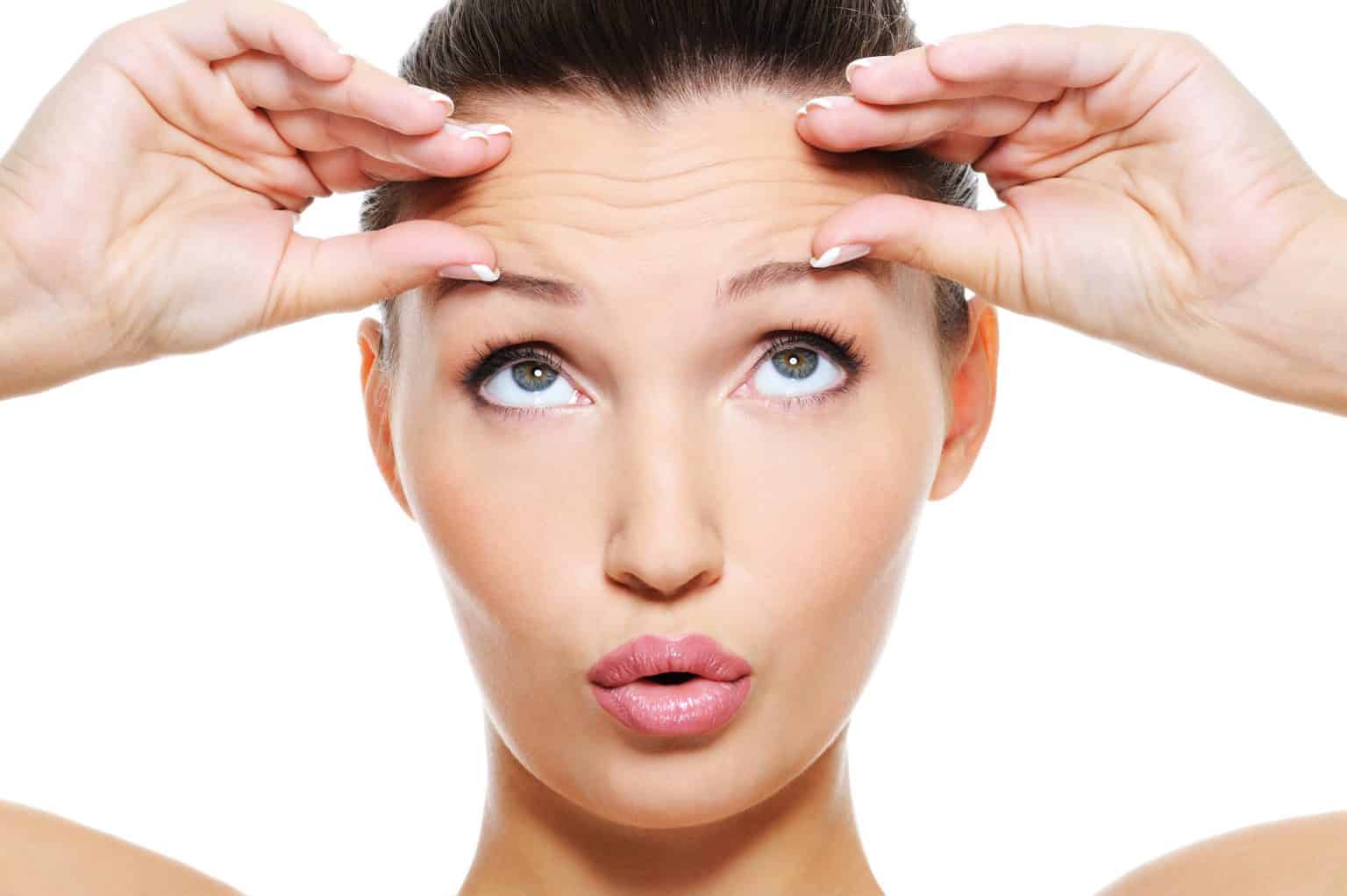Young woman pulling her forehead skin upward to show forehead lines against a white background