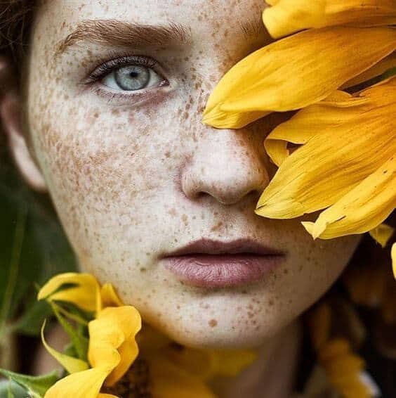 Close-up of a freckled young woman’s face partially covered by bright yellow sunflower petals