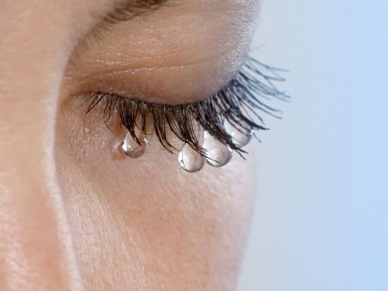 Close-up of a closed eye with tear droplets on the lower eyelashes against a light blue background