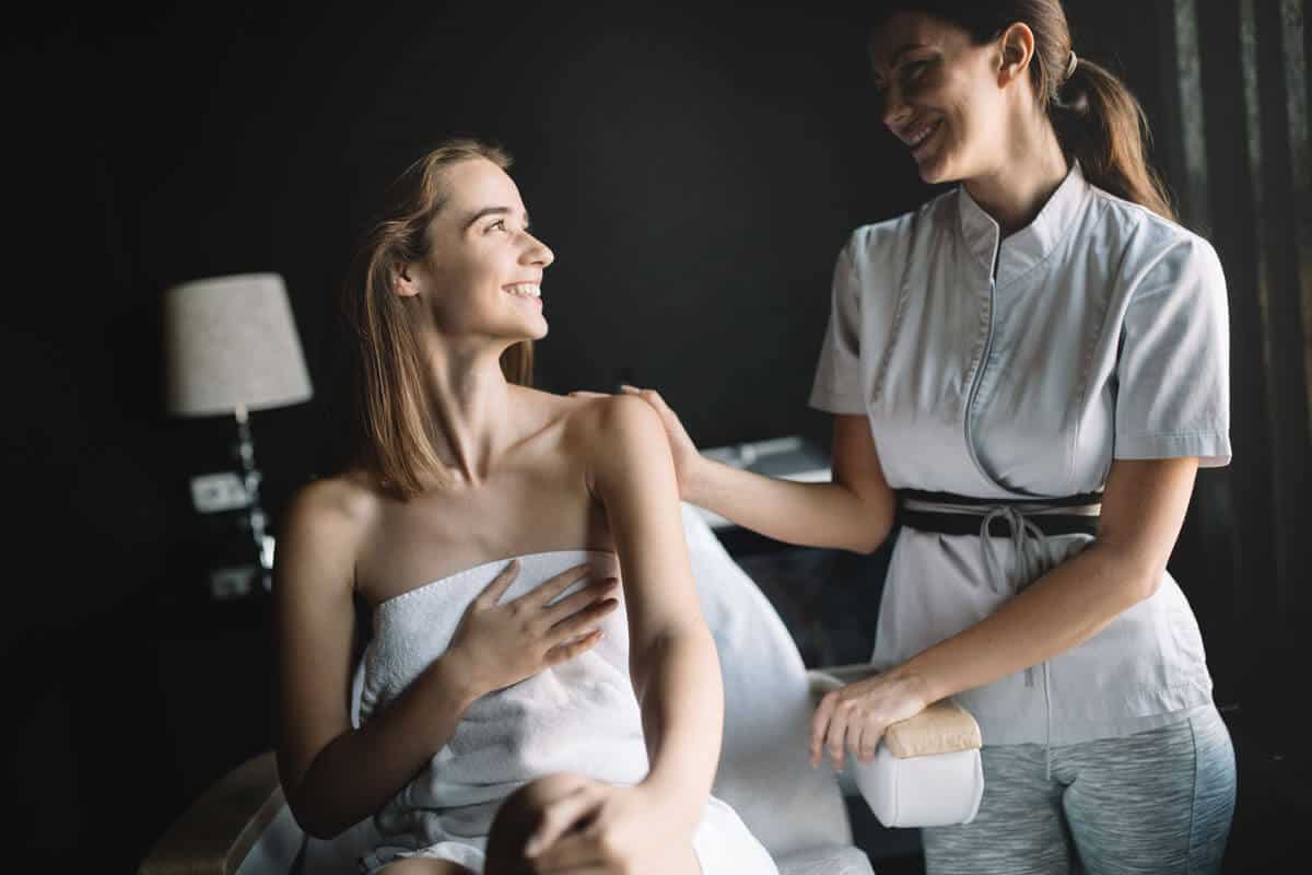 Smiling patient wrapped in a towel speaking with an esthetician in a white uniform in a dim treatment room