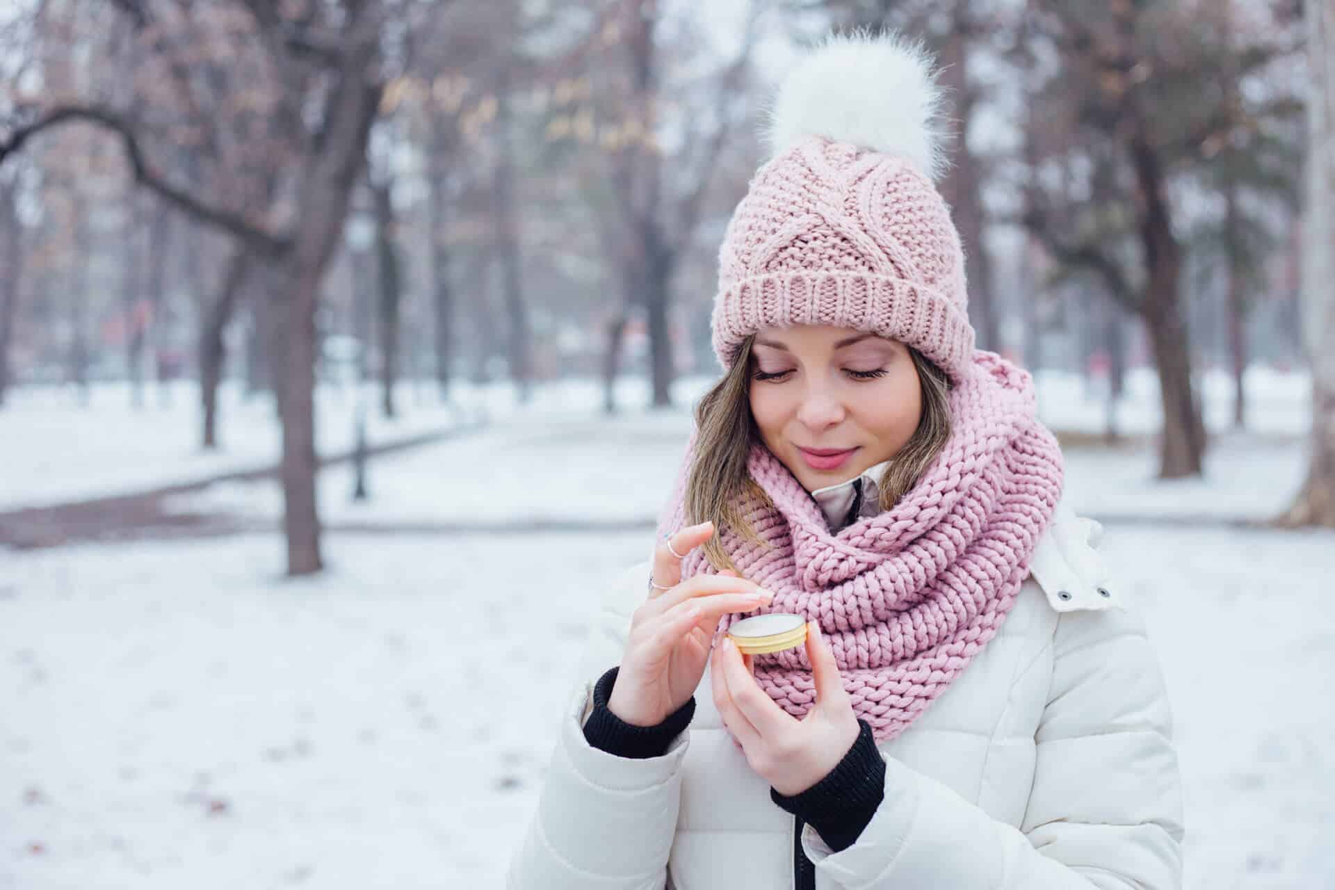 Woman applying moisturizer in a snowy park wearing pink knit hat and matching scarf