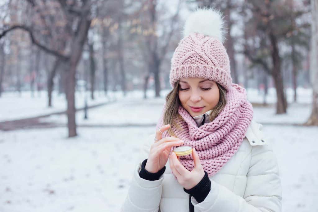 woman in a snowy park using a skin care treatment and wearing a pink hat, pink scarf and white coat